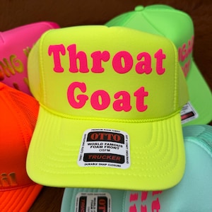 May include: A vibrant yellow trucker hat with the text "Throat Goat" in pink. The hat features a mesh back and a curved peak. The hat has an "OTTO" label on the front. Other hats in neon colours are visible.