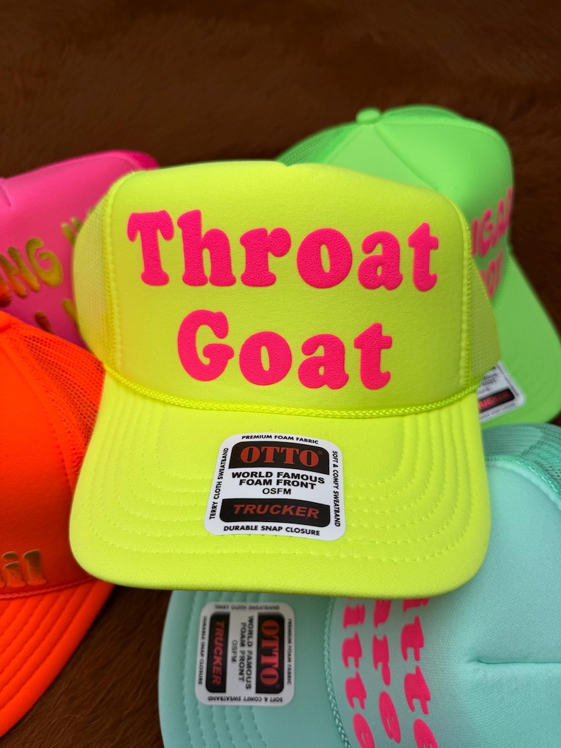 May include: A vibrant yellow trucker hat with the text "Throat Goat" in pink. The hat features a mesh back and a curved peak. The hat has an "OTTO" label on the front. Other hats in neon colours are visible.