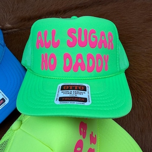 May include: A vibrant green trucker hat with the pink text "ALL SUGAR NO DADDY". The hat has a mesh back and a snap closure. The label reads "World Famous Foam Front" by Otto.