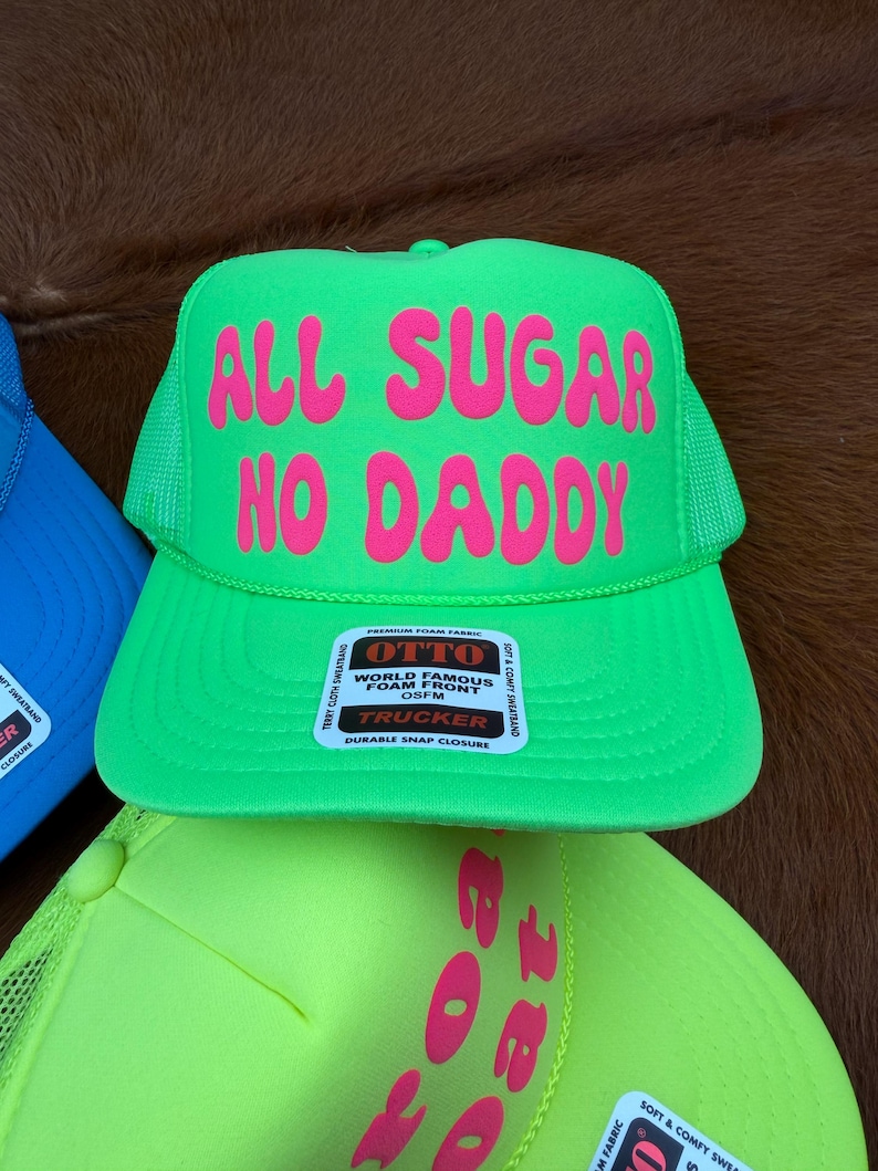 May include: A vibrant green trucker hat with the pink text "ALL SUGAR NO DADDY". The hat has a mesh back and a snap closure. The label reads "World Famous Foam Front" by Otto.