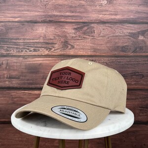 Op de afbeelding: Een beige baseballpet met een bruin leren patch waarop "YOUR TEXT / LOGO HERE" staat. De pet heeft een verstelbare band met de tekst "THE AUTHENTIC ADJUSTABLE YUPOONG SINCE 1974".