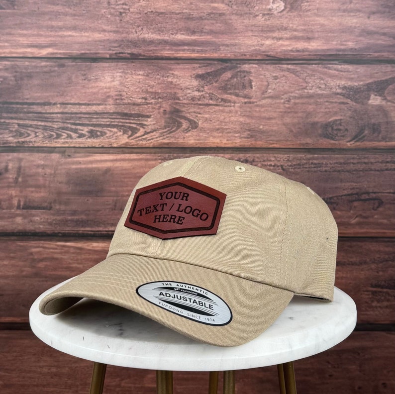 Op de afbeelding: Een beige baseballpet met een bruin leren patch waarop "YOUR TEXT / LOGO HERE" staat. De pet heeft een verstelbare band met de tekst "THE AUTHENTIC ADJUSTABLE YUPOONG SINCE 1974".