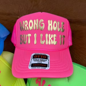 May include: A bright pink trucker hat with the gold text "WRONG HOLE BUT I LIKE IT". The hat features a mesh back and a curved peak. The label reads "OTTO" and "TRUCKER".