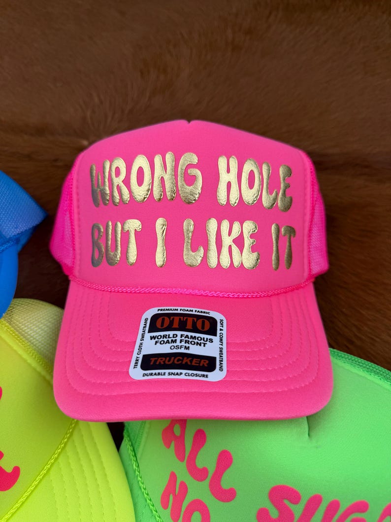 May include: A bright pink trucker hat with the gold text "WRONG HOLE BUT I LIKE IT". The hat features a mesh back and a curved peak. The label reads "OTTO" and "TRUCKER".