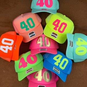 Puede incluir: Una colección de gorras de camionero en varios colores, incluyendo verde neón, rosa y azul. Cada gorra presenta el número "40" en un color contrastante en la parte delantera. Las gorras tienen una parte trasera de malla y una visera curva.