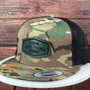 Op de afbeelding: Een camouflage trucker cap met een zwart mesh achterpaneel. De pet heeft een groen leren patch met de tekst "YOUR TEXT / LOGO HERE" erop.