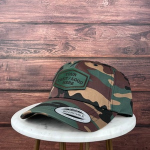 Op de afbeelding: Een camouflage baseballpet met een verstelbare band en een bruin leren patch met de tekst "YOUR TEXT/LOGO HERE".