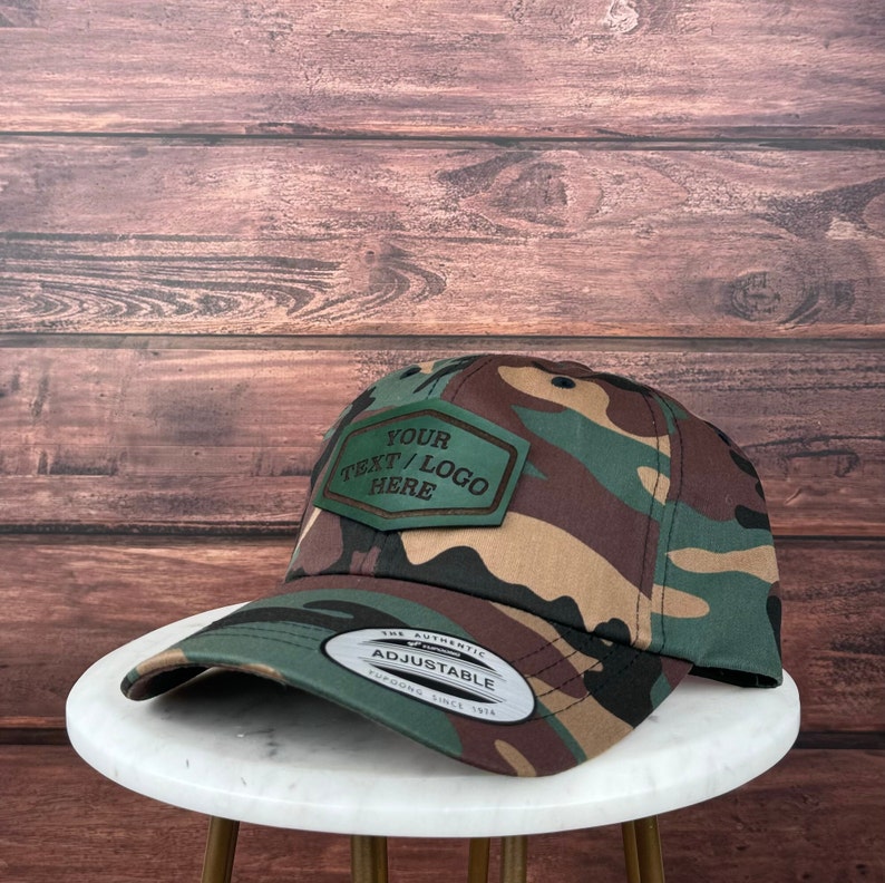 Op de afbeelding: Een camouflage baseballpet met een verstelbare band en een bruin leren patch met de tekst "YOUR TEXT/LOGO HERE".