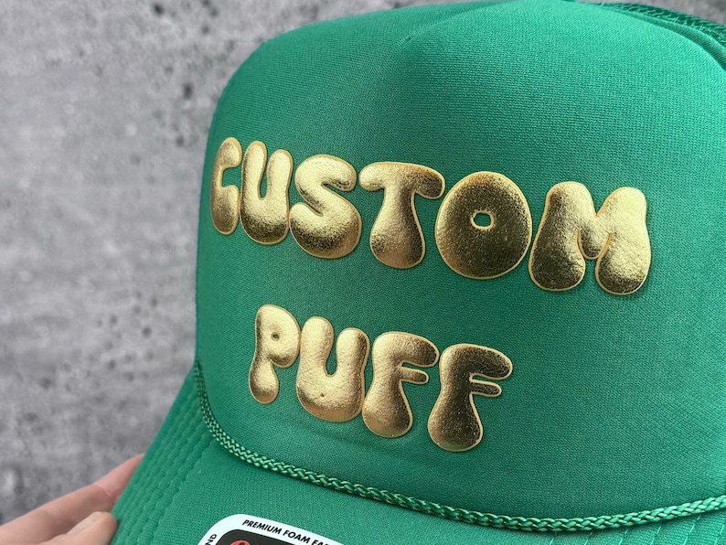 May include: Green trucker hat with the words "CUSTOM PUFF" in gold puffy letters. The hat has a mesh back and a curved brim. The text is in a bubble font.
