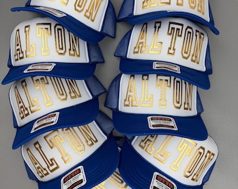 Könnte beinhalten: Eine Ansammlung von blau-weißen Trucker-Caps mit dem Namen "ALTON" in goldfarbenen Buchstaben auf der Vorderseite. Die Caps haben einen Mesh-Rücken und einen gebogenen Schirm. Jede Cap hat ein kleines Etikett.