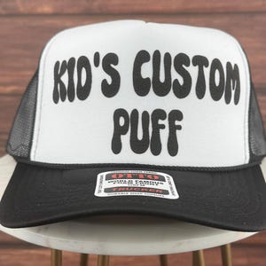 May include: A white trucker hat with a black mesh back. The front of the hat has black text that reads "Kid's Custom Puff" in a retro font.
