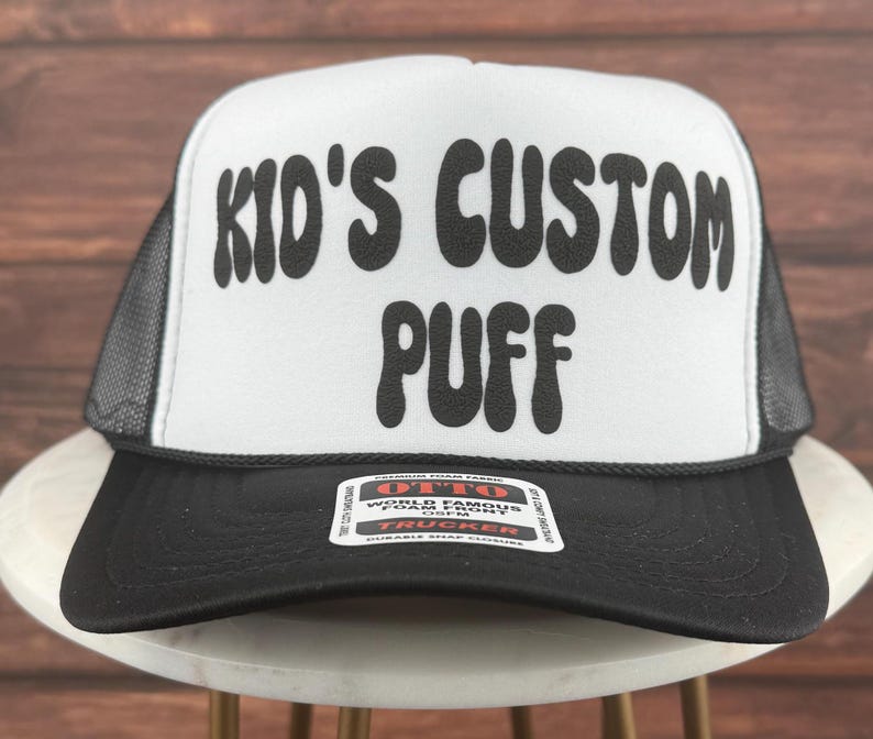 May include: A white trucker hat with a black mesh back. The front of the hat has black text that reads "Kid's Custom Puff" in a retro font.