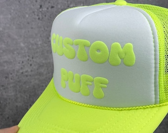 Op de afbeelding: Een neon groene en witte trucker cap met de tekst "CUSTOM PUFF" in neon groene bubbelletters.