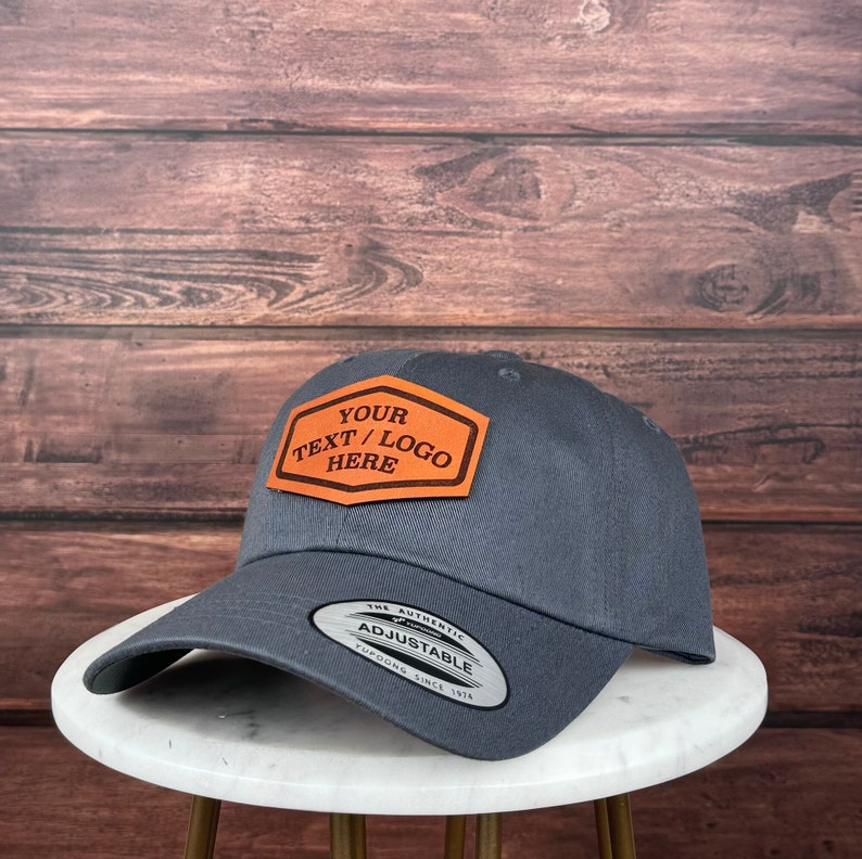 Op de afbeelding: Een donkergrijze baseballpet met een verstelbare band en een bruin leren patch met de tekst "YOUR TEXT/LOGO HERE".