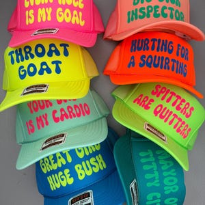 May include: A collection of trucker hats in various colors, including pink, yellow, orange, green, and blue. Each hat features bold, humorous text in contrasting colors. The hats have a mesh back and a foam front.
