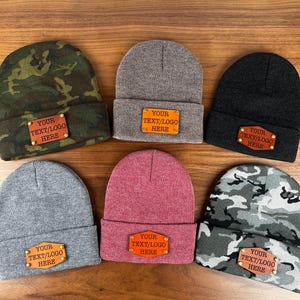 May include: Six different knit beanies with leather patches that say "YOUR TEXT/LOGO HERE". The beanies are in various colors and patterns, including gray, black, red, and camouflage.