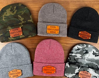 May include: Six different knit beanies with leather patches that say "YOUR TEXT/LOGO HERE". The beanies are in various colors and patterns, including gray, black, red, and camouflage.