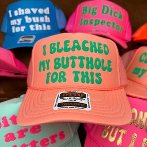 May include: A selection of trucker hats in shades of blue, pink, and orange. Each hat has a mesh back and features humorous text in contrasting colours. The text on the hats includes phrases like "I shaved my bush for this" and "I bleached my butthole for this."