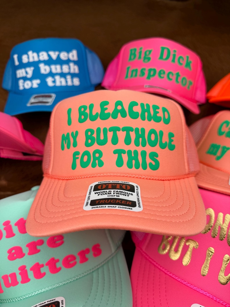 May include: A selection of trucker hats in shades of blue, pink, and orange. Each hat has a mesh back and features humorous text in contrasting colours. The text on the hats includes phrases like "I shaved my bush for this" and "I bleached my butthole for this."