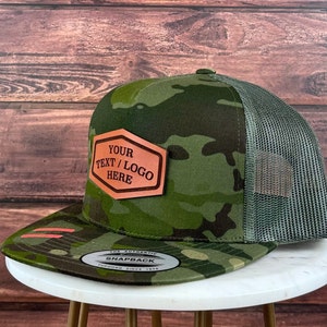 May include: A green camouflage trucker hat with a brown leather patch that says "YOUR TEXT/LOGO HERE".