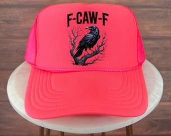 May include: A neon pink trucker hat with a black raven perched on a tree branch graphic. The hat features the text "F-CAW-F" in black block letters across the front. The hat has a mesh back and a curved brim.