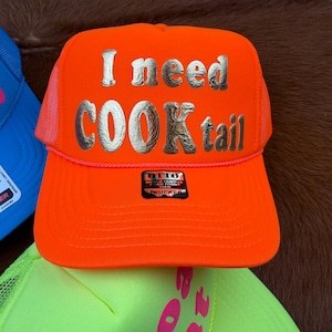May include: Vibrant orange trucker cap featuring the text "I need COOKtail" in silver. The cap has a mesh back and a curved peak. The brand name "OTTO" is displayed on a small label.