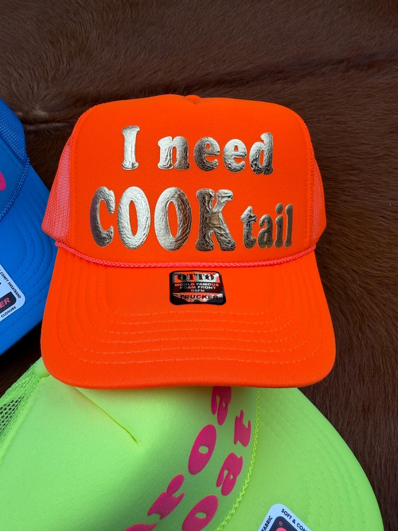 May include: Vibrant orange trucker cap featuring the text "I need COOKtail" in silver. The cap has a mesh back and a curved peak. The brand name "OTTO" is displayed on a small label.