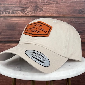 Op de afbeelding: Een beige baseballpet met een verstelbare band en een bruin leren patch met de tekst "YOUR TEXT/LOGO HERE".
