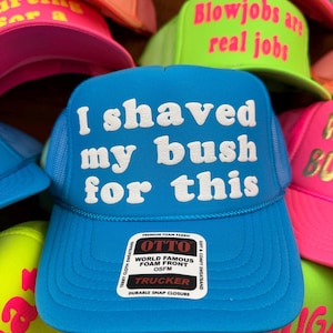 May include: A selection of brightly coloured trucker hats. The central blue hat has the text "I shaved my bush for this" in white. Other hats feature phrases such as "Blowjobs are real jobs" and "Big Dick Inspector".