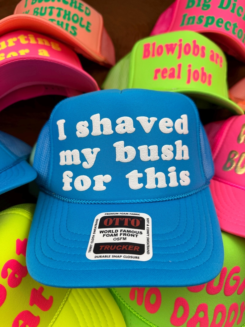 May include: A selection of brightly coloured trucker hats. The central blue hat has the text "I shaved my bush for this" in white. Other hats feature phrases such as "Blowjobs are real jobs" and "Big Dick Inspector".