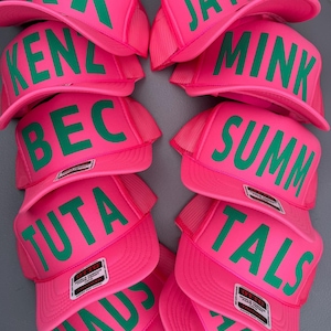 May include: A collection of pink trucker hats with green lettering. Each hat displays a different name or word, including "KK", "Jayme", "Kenz", "Mink", "Bec", "Summ", "Tuta", "Tals", "Maus", and "Karly". The hats have a mesh back and a foam front.