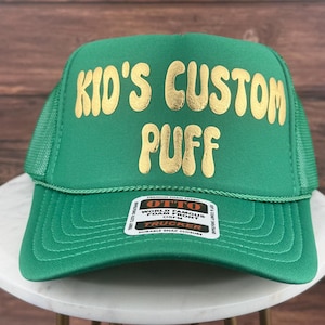May include: Green trucker hat with a gold embroidered design that says "Kid's Custom Puff".