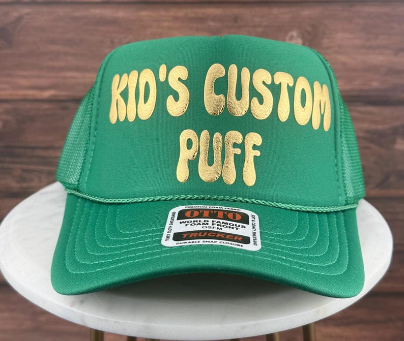 May include: Green trucker hat with a gold embroidered design that says "Kid's Custom Puff".