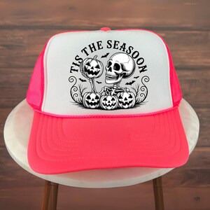 May include: A neon pink and white trucker hat with a Halloween graphic. The design includes a skeleton, pumpkins, and the text "Tis the Season". The hat has a mesh back and a curved peak.