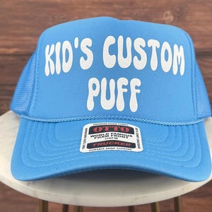 May include: A light blue trucker hat with a white mesh back. The front of the hat has white text that says "Kid's Custom Puff". The hat has a red label that says "Otto World Famous Foam Front OS/FM Trucker Durable Snap Closure".