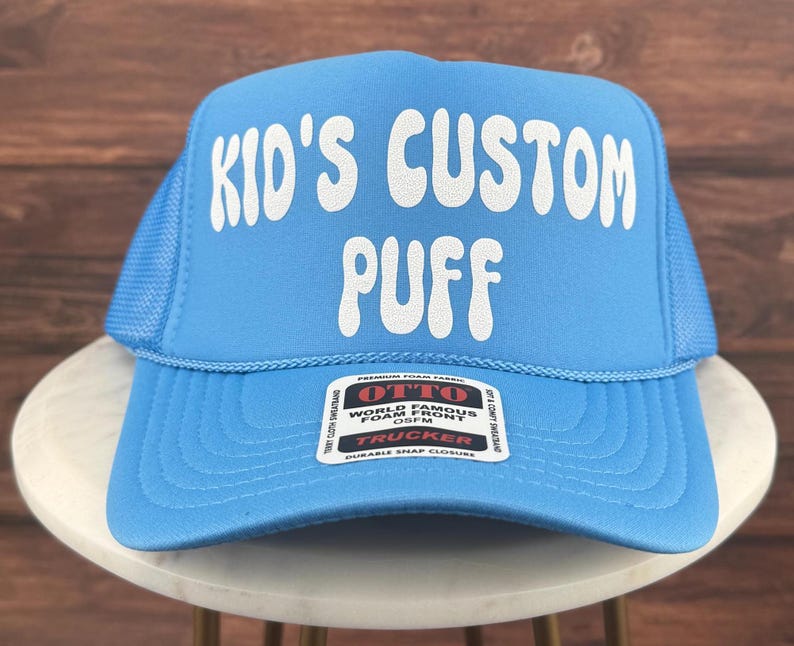 May include: A light blue trucker hat with a white mesh back. The front of the hat has white text that says "Kid's Custom Puff". The hat has a red label that says "Otto World Famous Foam Front OS/FM Trucker Durable Snap Closure".