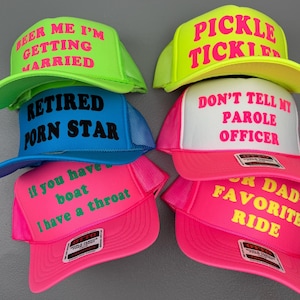 May include: A collection of six foam trucker hats in neon green, bright blue, hot pink, and white. Each hat features bold text: BEER ME I'M GETTING MARRIED, PICKLE TICKLED, RETIRED PORN STAR, DON'T TELL MY PAROLE OFFICER, IF YOU HAVE A BOAT I HAVE A THROAT, and YOUR DAD'S FAVORITE RIDE.