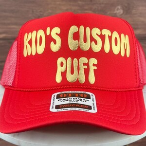 May include: Red trucker hat with a gold embroidered design that says "Kid's Custom Puff". The hat has a black label with white text that says "Otto World Famous Foam Front OSFM Trucker Durable Snap Closure".