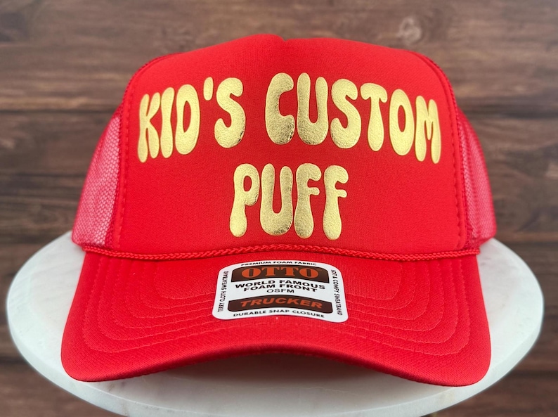 May include: Red trucker hat with a gold embroidered design that says "Kid's Custom Puff". The hat has a black label with white text that says "Otto World Famous Foam Front OSFM Trucker Durable Snap Closure".