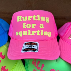 May include: A bright pink trucker hat with the words "Hurting for a squirting" in yellow. The hat features a mesh back and a snap closure. Other hats in different colours are also visible.