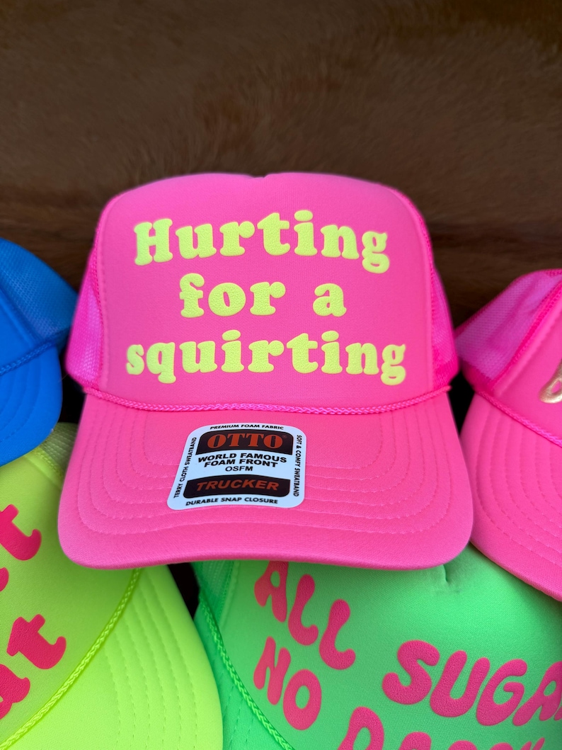 May include: A bright pink trucker hat with the words "Hurting for a squirting" in yellow. The hat features a mesh back and a snap closure. Other hats in different colours are also visible.
