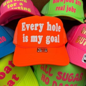May include: A selection of neon trucker caps, each displaying bold text. The central orange cap reads "Every hole is my goal" in white. Other caps feature phrases like "Hurting for a rating" and "Blowjobs are real jobs" in pink and green.