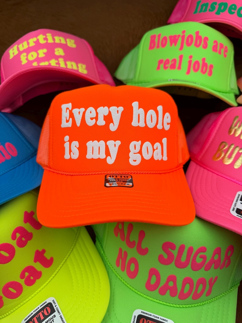 May include: A selection of neon trucker caps, each displaying bold text. The central orange cap reads "Every hole is my goal" in white. Other caps feature phrases like "Hurting for a rating" and "Blowjobs are real jobs" in pink and green.