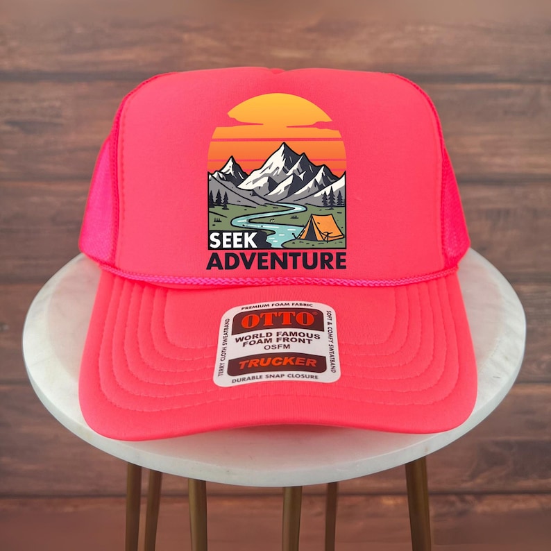 Peut inclure: Casquette de camionneur rose vif avec un dos en filet et une visi&egrave;re incurv&eacute;e. Le panneau avant pr&eacute;sente un graphique de montagne avec le texte "SEEK ADVENTURE". La casquette a une fermeture &agrave; pression et est en tissu mousse.
