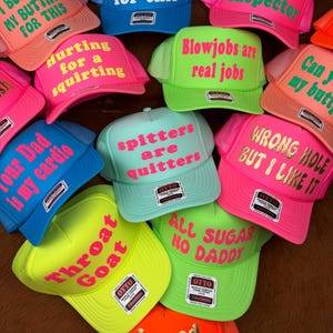 Könnte beinhalten: Eine Sammlung von Trucker-Caps in verschiedenen leuchtenden Farben, darunter Pink, Blau, Grün und Orange. Jede Kappe hat einen humorvollen Text in kontrastierenden Farben. Die Kappen haben einen Mesh-Rücken und einen gebogenen Schirm.