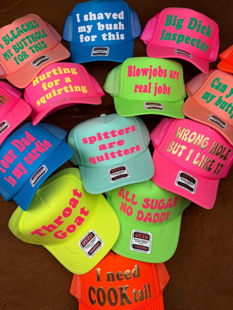 May include: A selection of brightly coloured trucker hats, including pink, blue, green, and orange. Each hat displays bold text in contrasting colours, with phrases such as "I shaved my bush for this" and "Throat Goat".