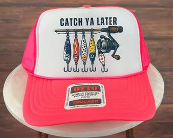 Puede incluir: Gorra de camionero con panel frontal blanco y visera y parte trasera de malla rosa brillante. La gorra presenta el texto "CATCH YA LATER" sobre una caña de pescar con señuelos. Cierre a presión.