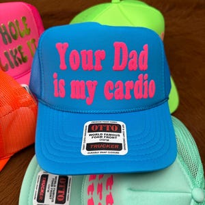 May include: Several trucker hats in vibrant colours are displayed. A blue hat has the pink text "Your Dad is my cardio". Other hats include the phrases "HOLE LIKE IT". The hats have a mesh back.