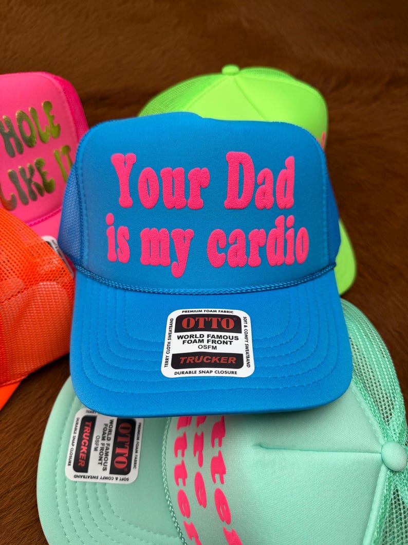 May include: Several trucker hats in vibrant colours are displayed. A blue hat has the pink text "Your Dad is my cardio". Other hats include the phrases "HOLE LIKE IT". The hats have a mesh back.