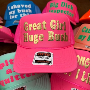 May include: A selection of trucker hats in vibrant colours like pink, blue, and green. The hats display text in gold and white, including phrases like "Great Girl Huge Bush" and "I shaved my bush for the". Each hat has a foam front and a snap closure.